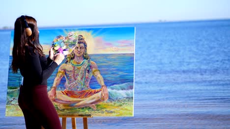 summer, near river, on beach. at sunrise, a beautiful woman artist holds a brush, palette in her hands, draws a picture. she paints on canvas god shiva. listening to music on headphones