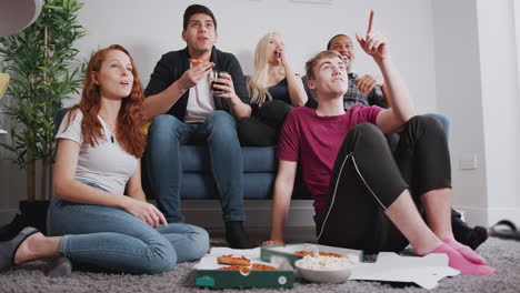 group of college students in shared house watching tv and eating pizza