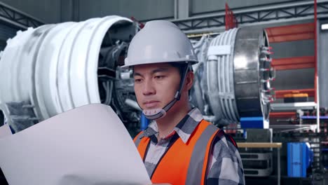 close up side view of asian male engineer with safety helmet looking at blueprint in his hands and looking around with airplane engine maintenance conducted, analytics checking the turbine