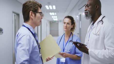 Diverse-group-of-a-female-and-two-male-doctors-talking-in-hospital-corridor-holding-tablet-and-files