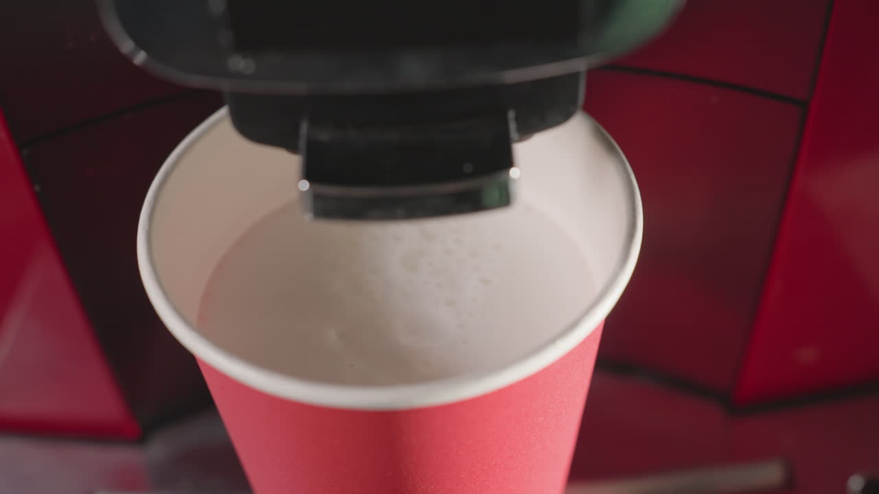 Close up of coffee machine dispensing foamy milk into red paper cup. Soft foam swirling in cup, ideal for cafe, coffee preparation, barista art, or milk-based beverages