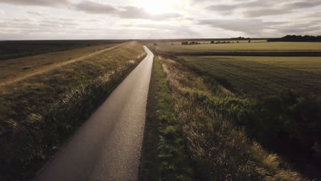 fast-drone-flight-backwards-above-an-empty-dyke-road,-back-light,-scenic-landscape,-sunset,-sky-with-scenic-clouds