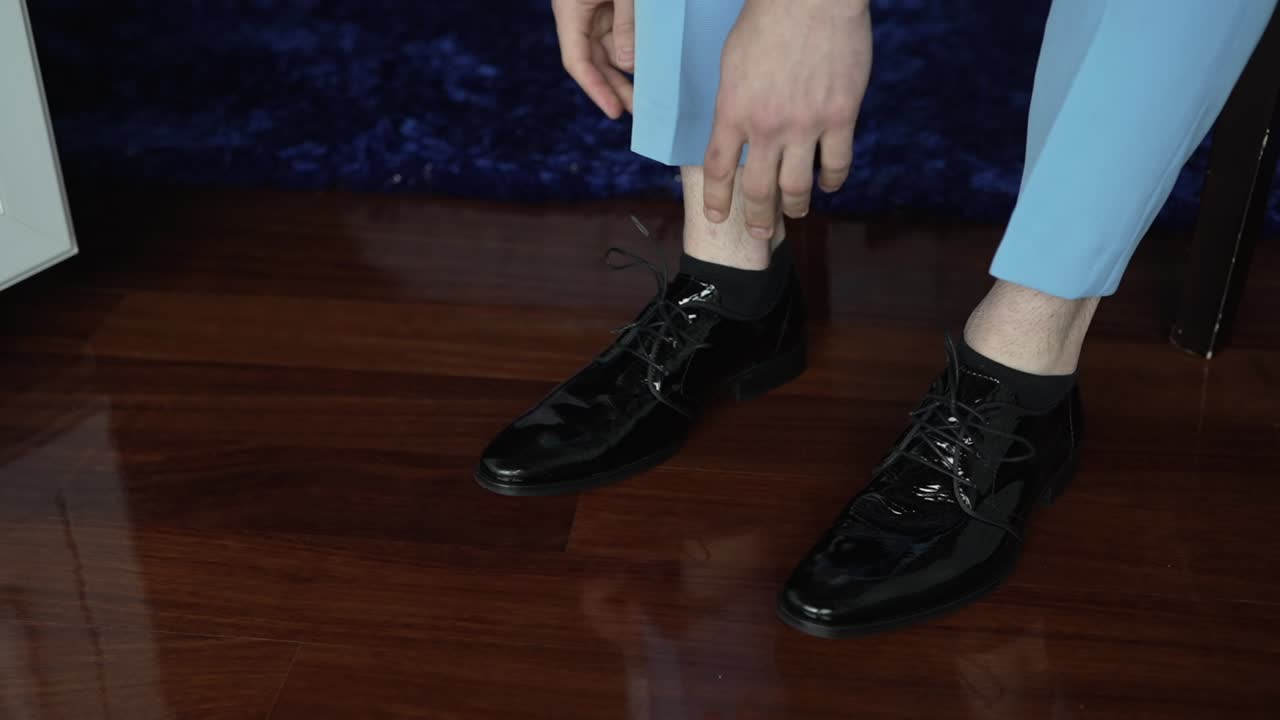 Close up of groom in light blue pants tying shiny black dress shoes on wooden floor