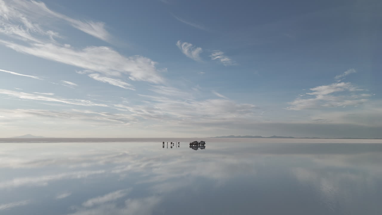 Drone shot flying above the mirror salt flats in Bolivia Uyuni on acloudy day with the visible reflection going towards a tourist truck with people around it LOG