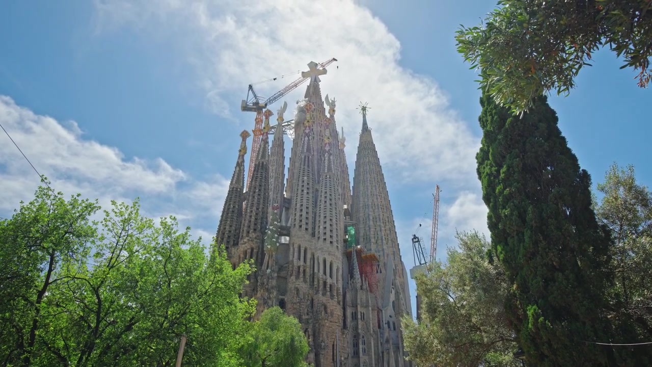 La Basilica della Sagrada Familia che si erge tra alberi rigogliosi a Barcellona, Spagna, con l'iconica architettura di Gaudí incorniciata dal verde fogliame sotto un limpido cielo estivo.