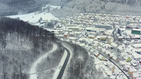 Winter-Von-Oben---Drohnenkamera-Verfolgt-Ein-Auto,-Das-An-Einer-Kurvenstraße-Und-Einer-Stadt-Neben-Der-Straße-Durch-Eine-Verschneite-Landschaft-Fährt