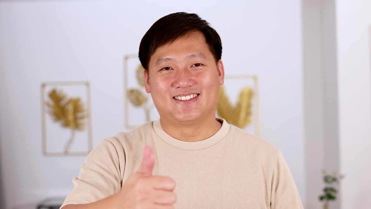 Asian man smiles and gives a thumbs up gesture in a well-lit, modern living room with minimal decor, captured in a steady medium shot