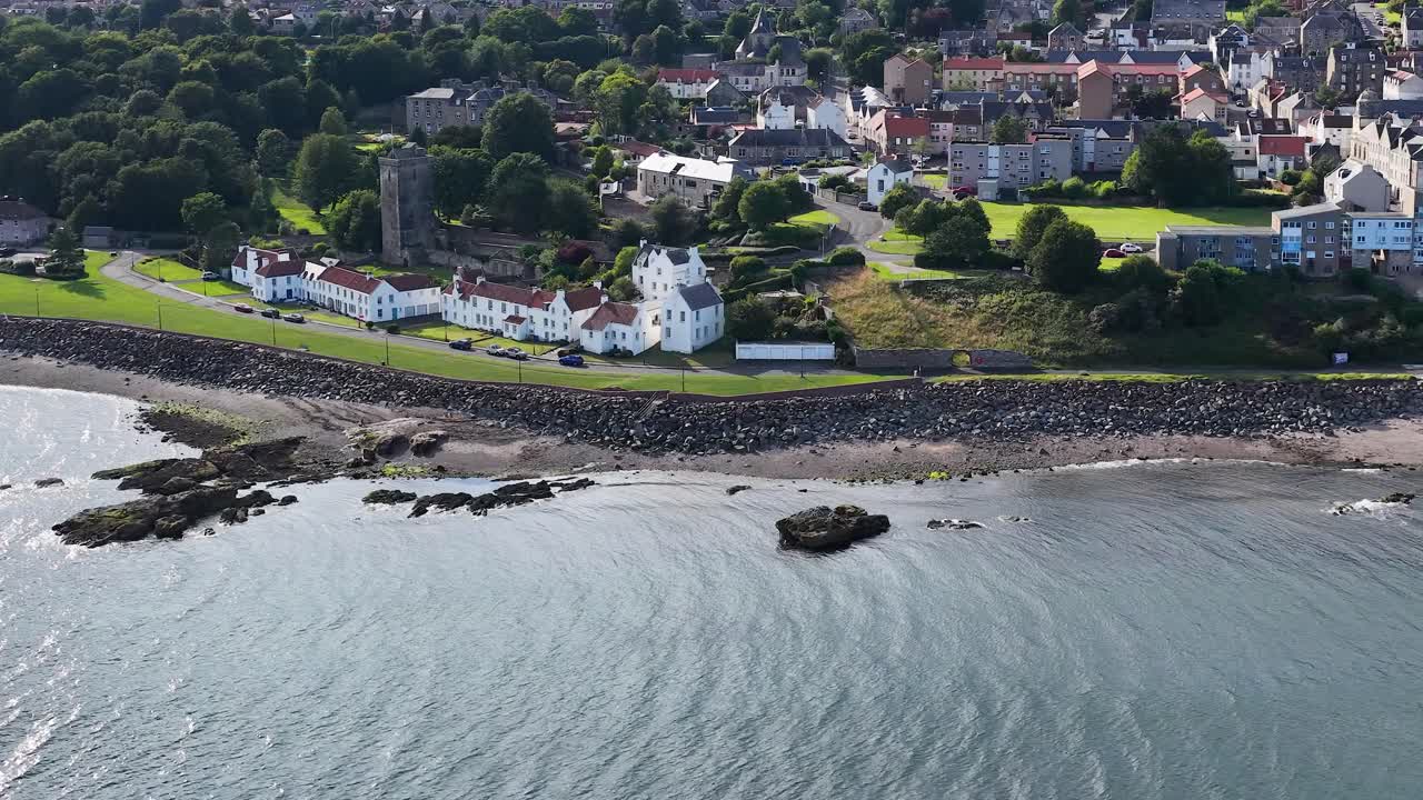 Drone pans above seaside town, revealing historic buildings, shoreline, and harbor in bright daylight