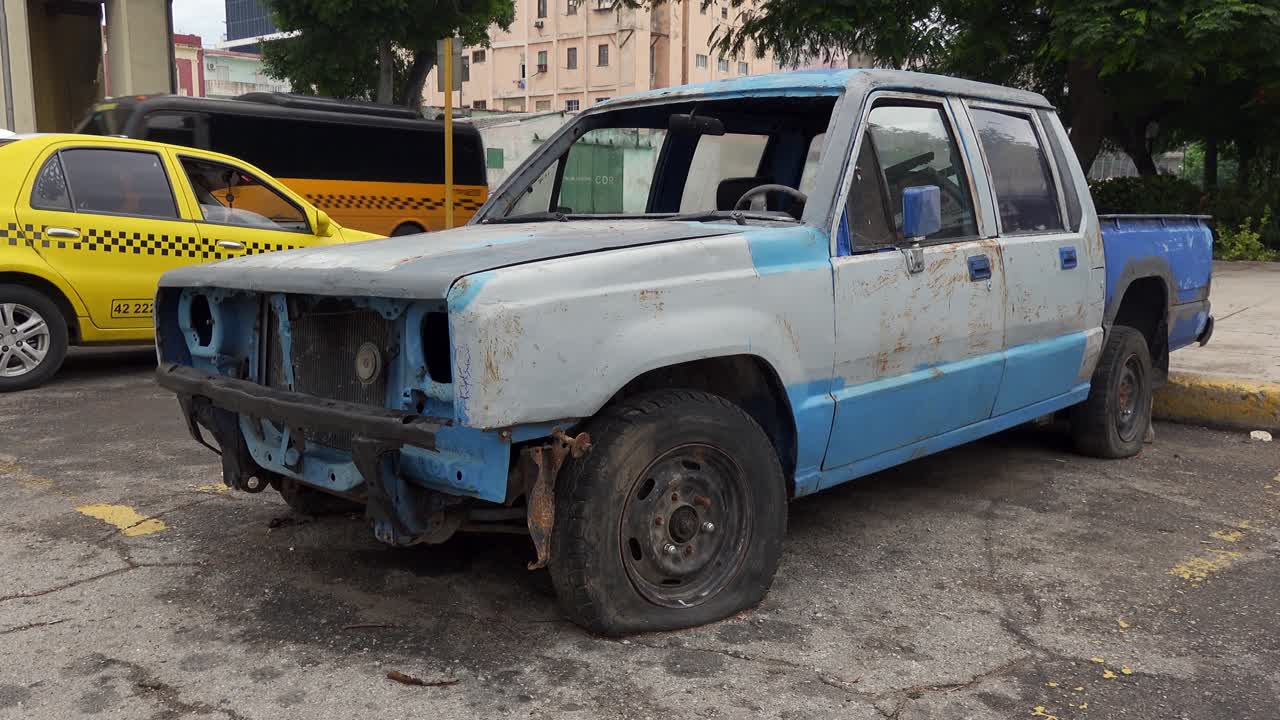 A rusty and dilapidated pickup truck with a flat tire parked on a city street next to a yellow taxi.