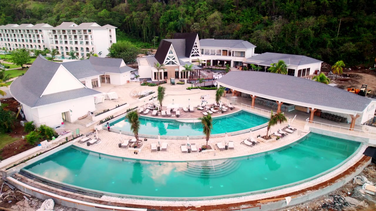 Una ripresa aerea con drone dall'alto che mostra la disposizione architettonica di un resort di lusso, evidenziando il complesso di piscine a più livelli, gli edifici per gli ospiti circostanti e i giardini paesaggistici.