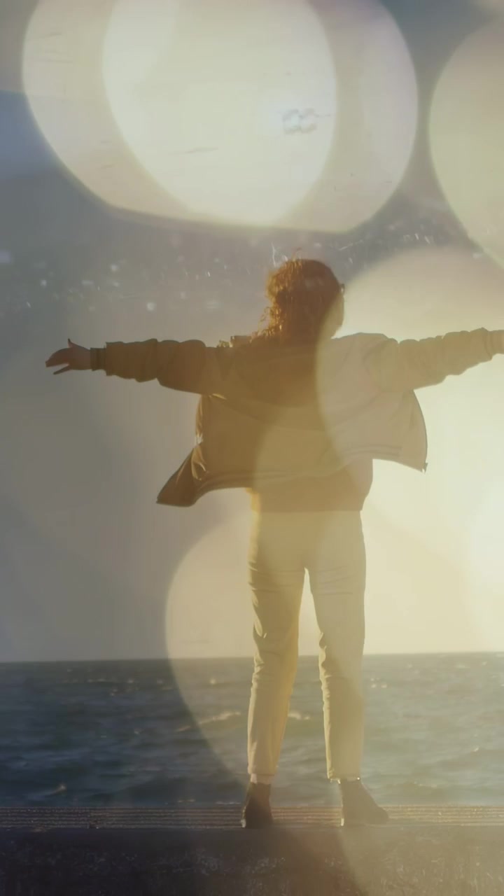 Video verticale: Donna che allarga le braccia al vento marino che le solleva il cappotto, con effetto bokeh attraverso l'ombra, per una pubblicità di viaggi.