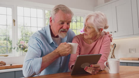 Retired-Senior-Couple-Sitting-In-Kitchen-At-Home-Drinking-Coffee-And-Using-Digital-Tablet