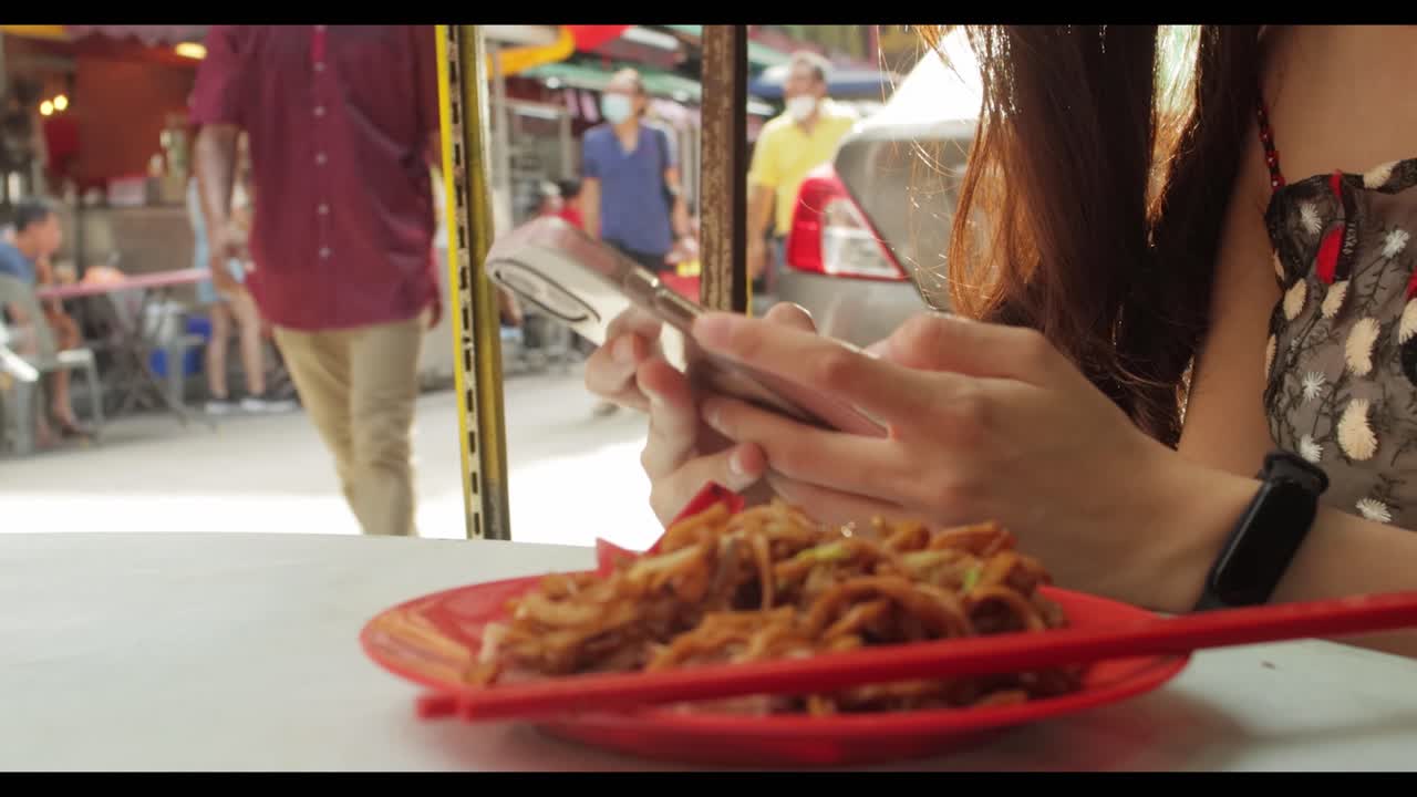 chica guapa asiática enviando mensajes de texto por teléfono con comida en la mesa mientras la gente pasa por el barrio chino de malasia durante el día