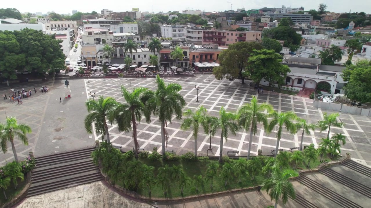 Ripresa aerea con drone in movimento di Plaza España che mostra lo storico Alcázar de Colón, l'architettura in pietra, le palme reali e la griglia urbana della città a Santo Domingo.
