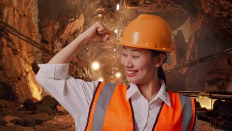 close up of asian female engineer with safety helmet flexing her bicep and smiling to camera in underground mine tunnel