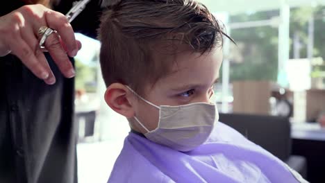 El-Peluquero-Corta-El-Pelo-De-Un-Niño-Caucásico,-Con-Una-Máscara-Facial,-Toma-De-Cerca