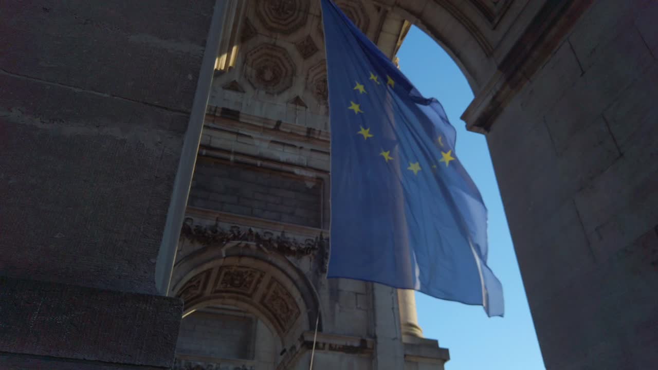 Bandiera dell'UE che sventola sotto un arco con ombre profonde e cielo sereno.