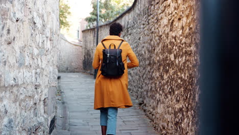 Back-view-of-young-black-woman-wearing-a-yellow-pea-coat-putting-smartphone-in-the-back-pocket-of-her-jeans-and-walking-away-from-camera-down-a-narrow-alleyway-between-stone-walls,-selective-focus