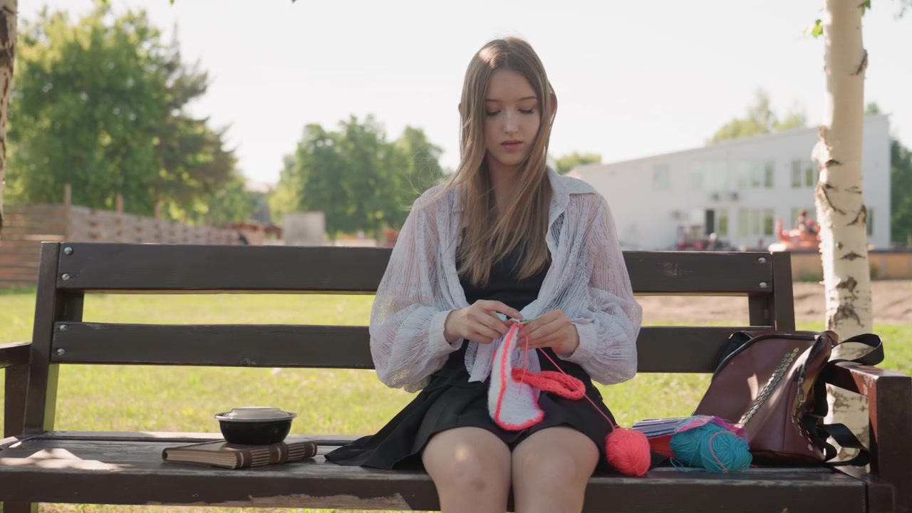 屋外の公園のベンチに座って赤い毛糸で編み物をする若い女性のクローズアップ。女性は白いブラウスと黒いスカートを着用し、明るい日差しの下、緑の木々に囲まれている。木製の