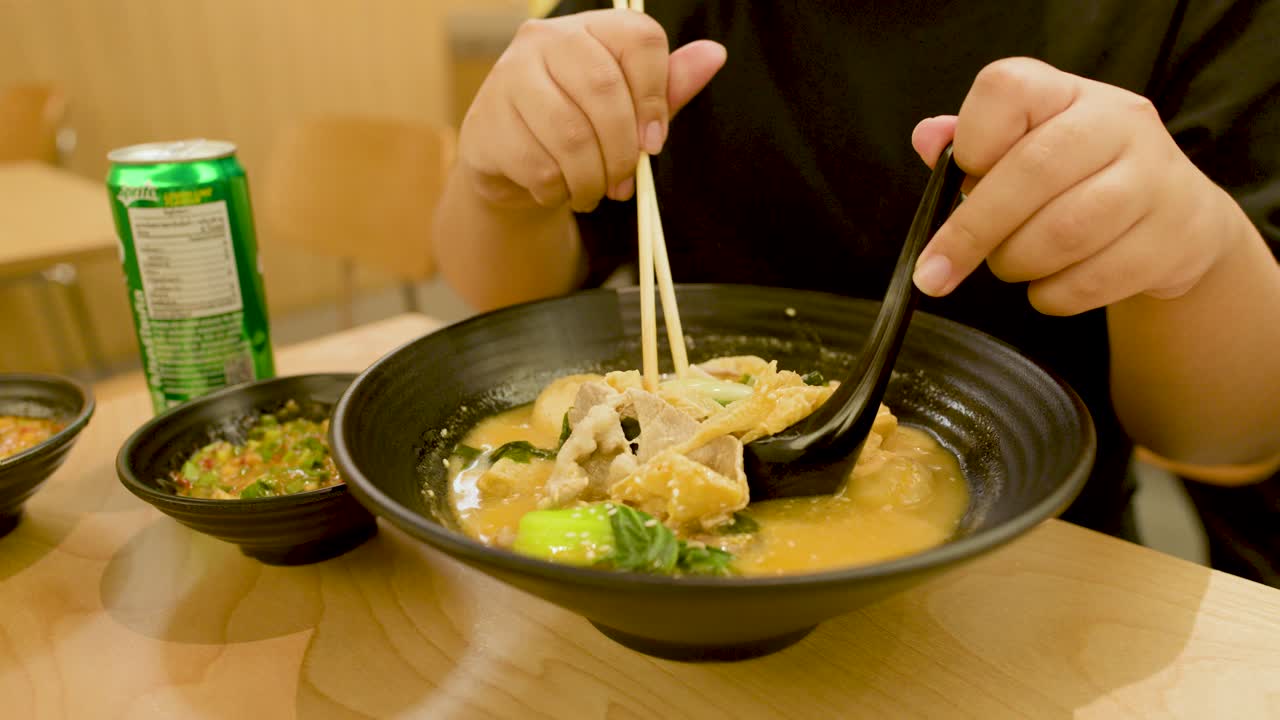 Person uses chopsticks and spoon to eat hot noodle soup in bright, casual dining setting