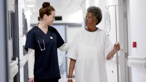 happy diverse female doctor walking with senior woman with drip in hospital corridor, slow motion