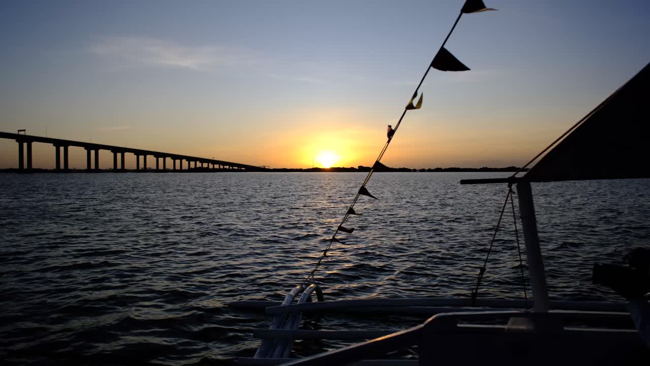 A serene Cebu sunset viewed from a traditional boat, with fluttering flags and a long bridge stretching across calm waters, creating a picturesque golden-hour scene.
