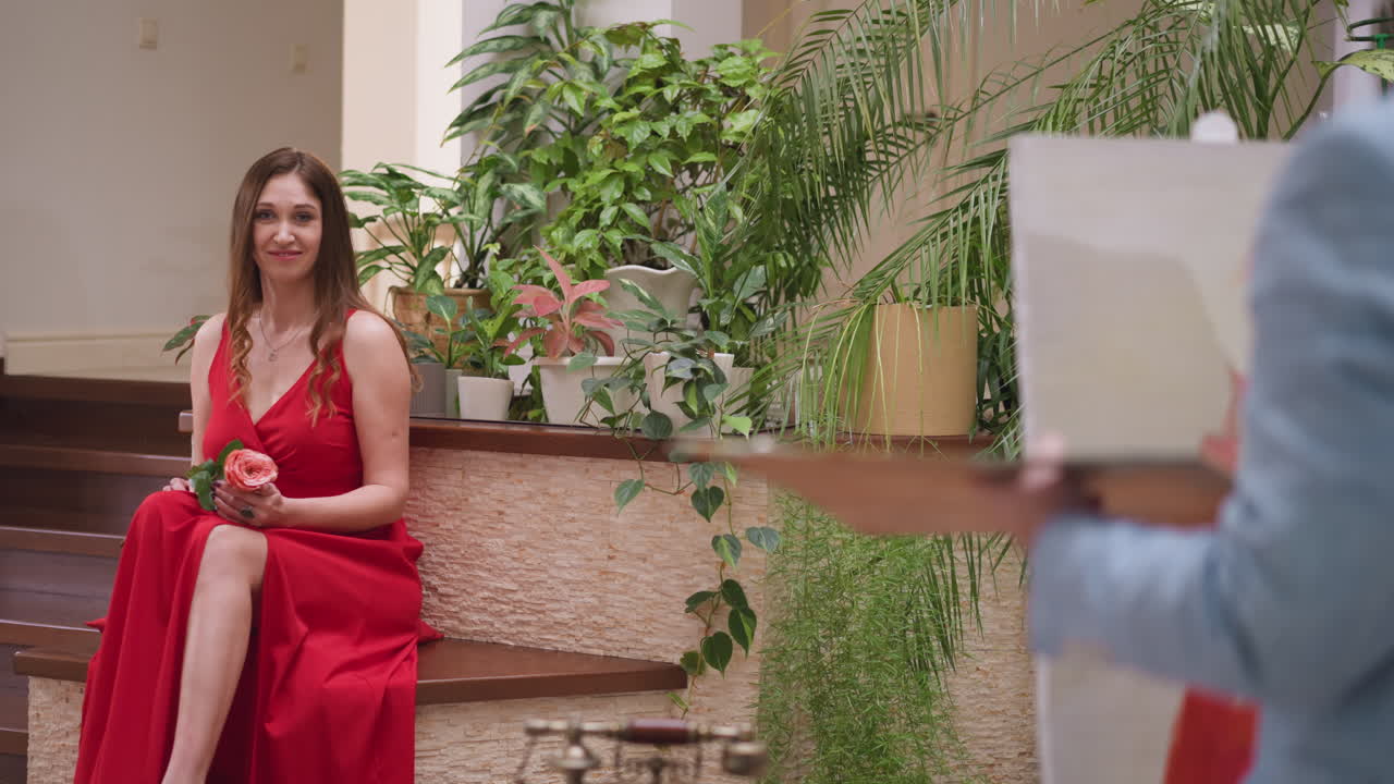 손에 장미를 들고 있는 여자를 그리는 예술가. 여성 모델은 예술 가게의 활기찬 분위기 속에서 화가의 상상력을 불러일으킨다. 매력적인 매력을 발산한다.