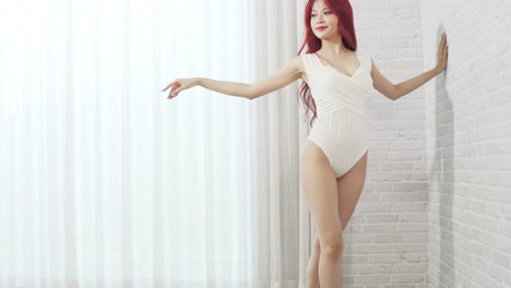 beautiful asian ballerina woman in white bodysuit long red hair wearing ballet shoes practicing posing ballet dance in the white room with wall and curtain background