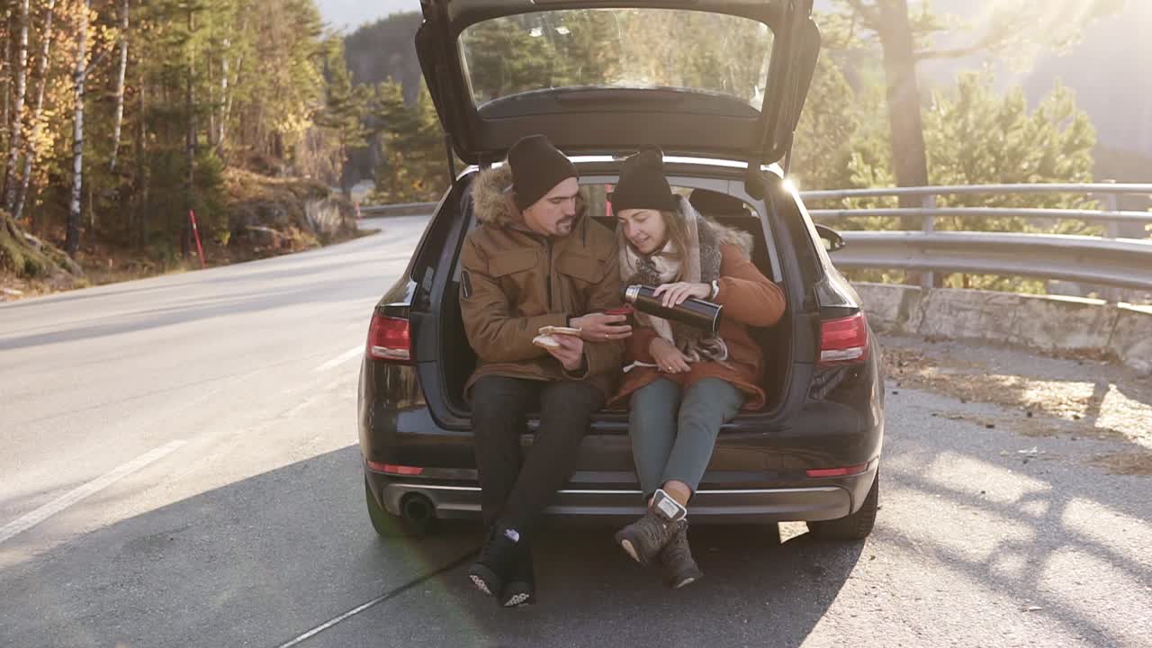 una pareja joven y amorosa bebe té caliente de un termo sentado en el baúl del auto. feliz pareja caucásica tomando un café durante un viaje por carretera en el campo, bebiendo y comiendo sándwiches. admirando la naturaleza