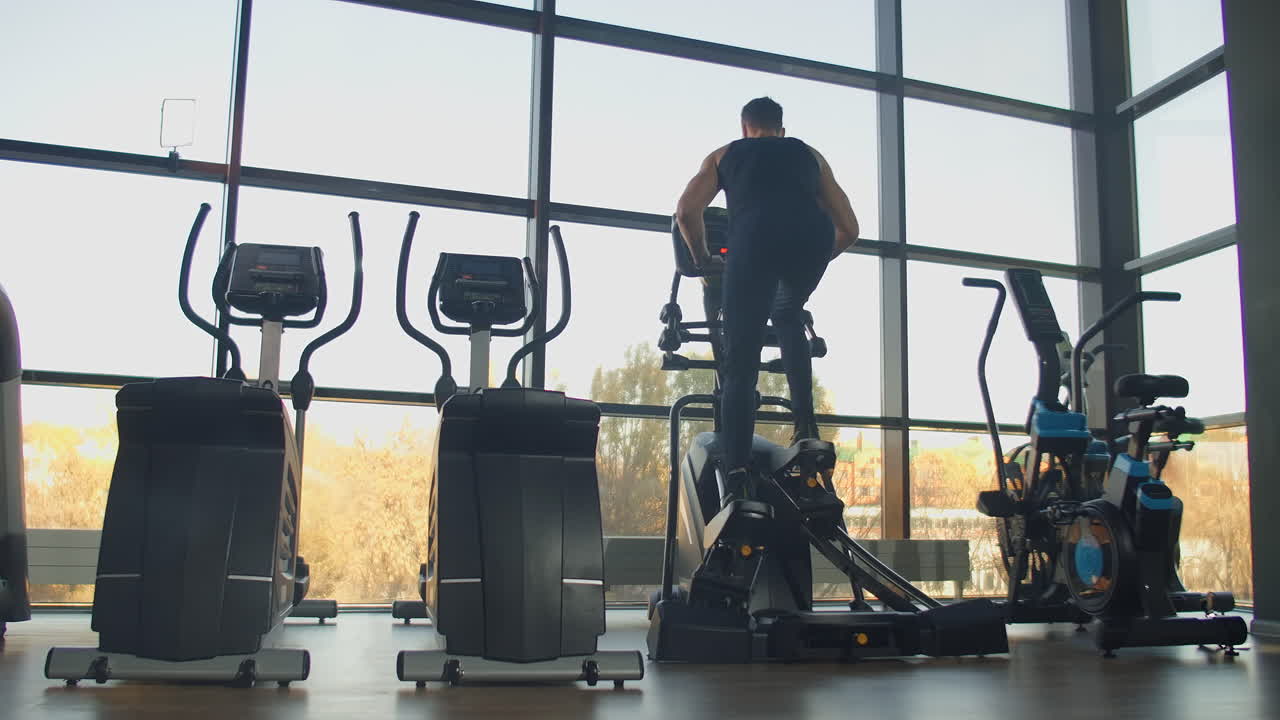 Back view a man in the cardio area of the gym trains on an elliptical trainer.
