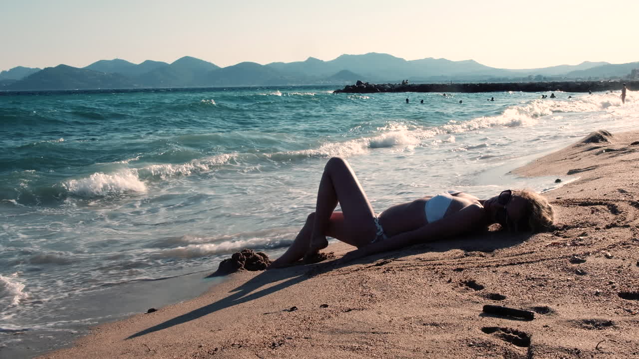 A young woman in a white bikini enjoys the sun on a sandy beach while a cruise ship sails in the distance