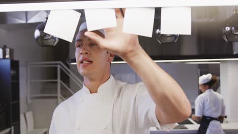 cocinero caucásico que trabaja en la cocina de un restaurante comprobando pedidos, con colegas que trabajan