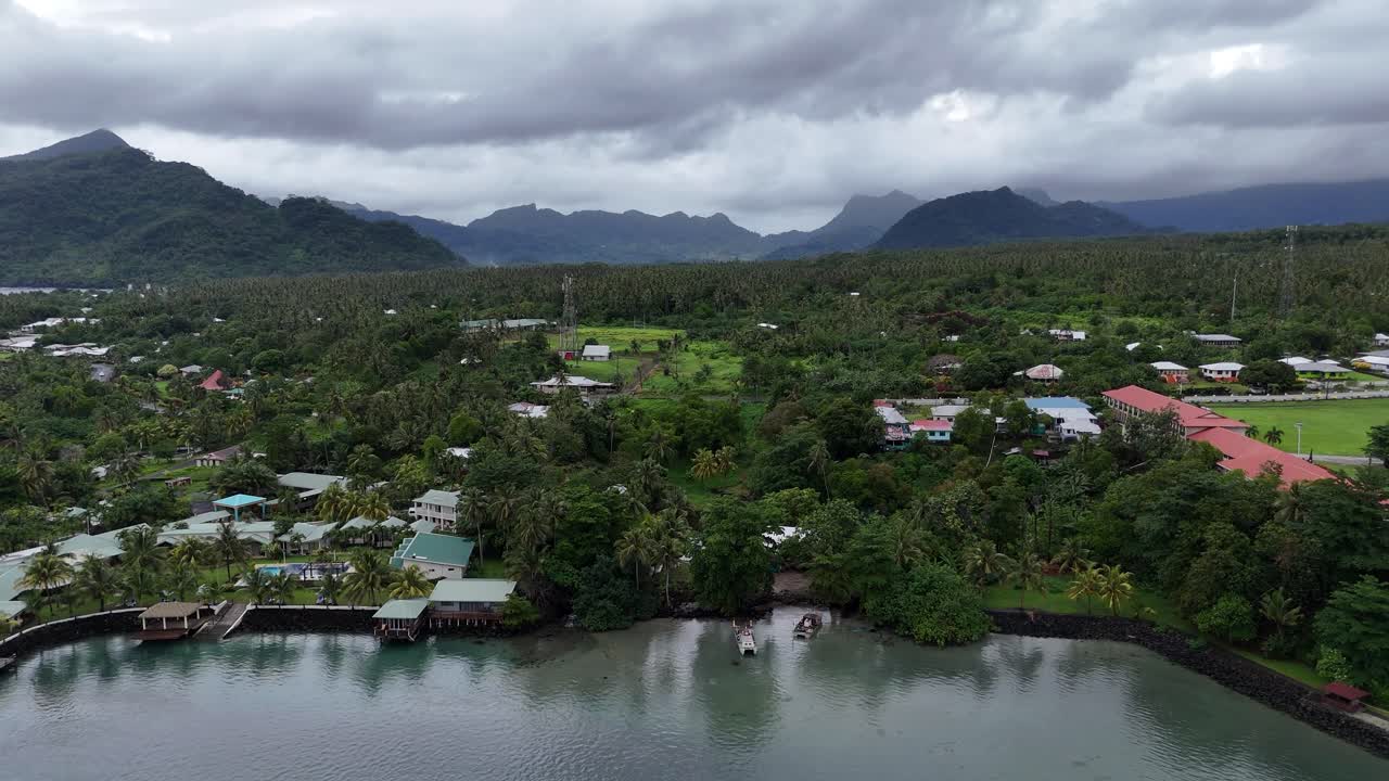 Diese Drohnenaufnahme mit Blick auf eine friedliche Küstensiedlung in Samoa fängt die traditionellen Häuser, das üppige Grün und die majestätischen Vulkane unter einem sanften Abendhimmel ein