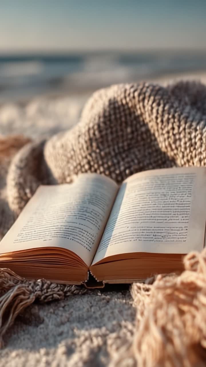 Una scena tranquilla di un libro aperto che riposa su sabbia soffice, circondato da una coperta accogliente e testurizzata, mentre dolci onde indugiano sullo sfondo, invitando a momenti di relax e riflessione.