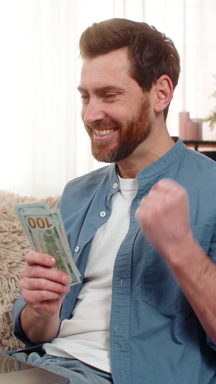 Hombre sosteniendo billetes de dólar en las manos, mirando pensativamente la pantalla de su laptop y decidiendo cómo gastar el dinero.