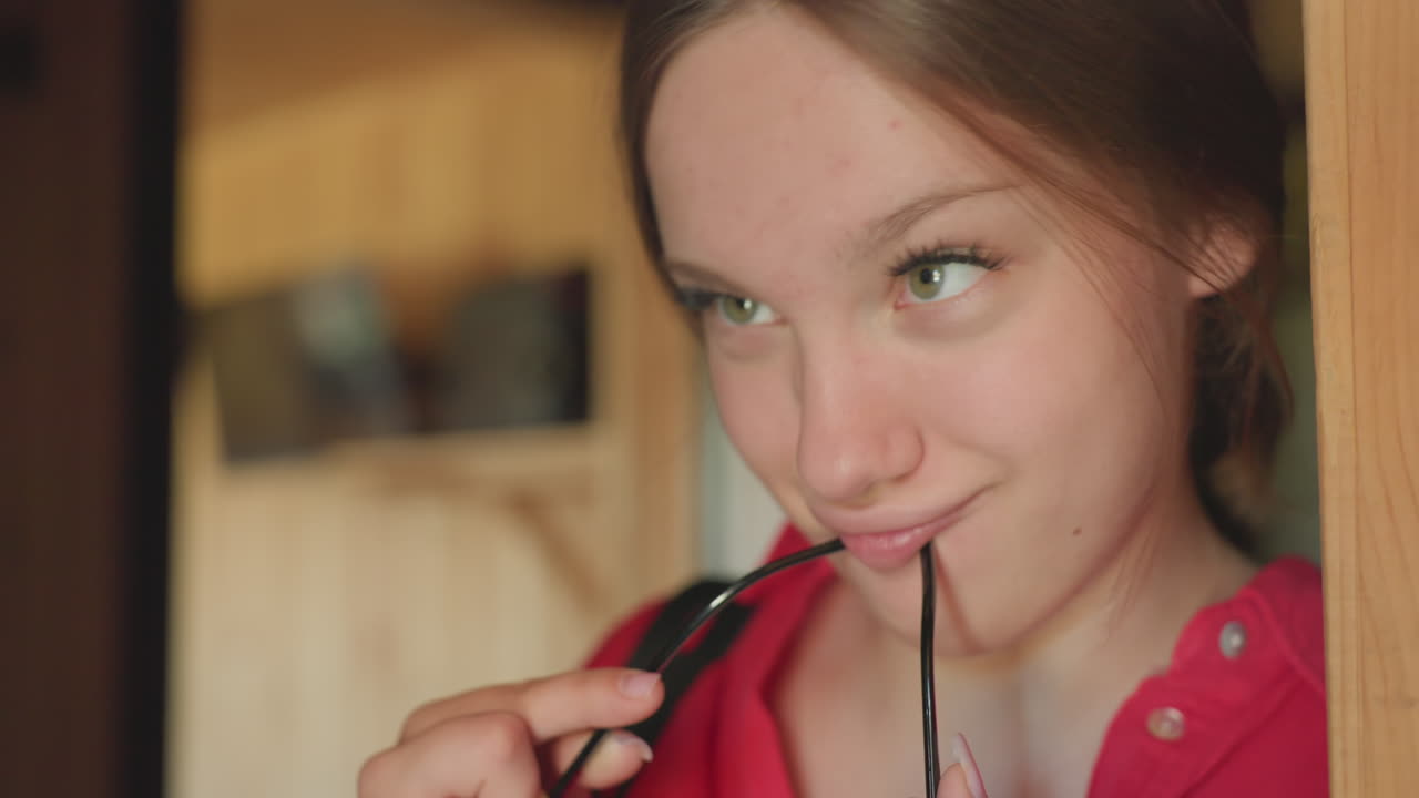 Young woman leaning on wooden doorframe while playfully making funny facial expression and holding glasses in mouth, expressing lighthearted attitude and casual mood in warm indoor environment