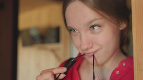 young woman leaning on wooden doorframe while playfully making funny facial expression and holding glasses in mouth, expressing lighthearted attitude and casual mood in warm indoor environment