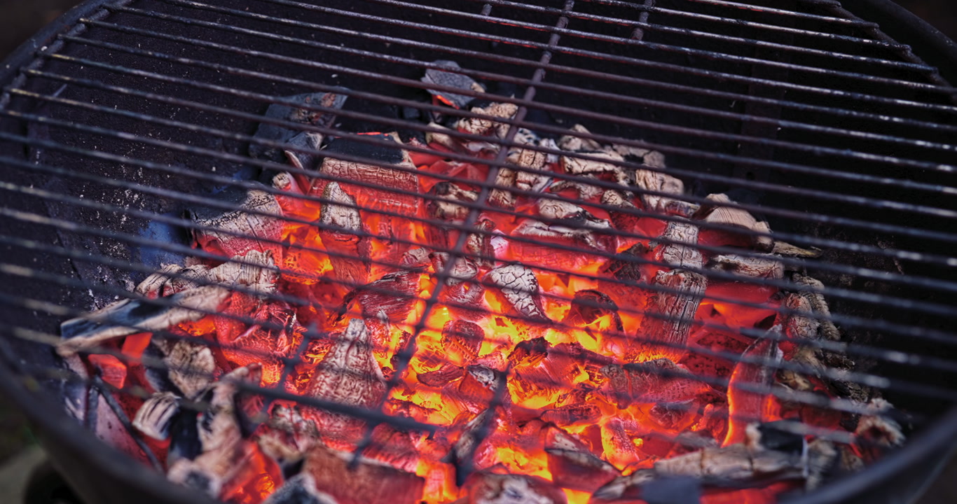 A smooth side glide passes over red hot coals beneath a worn grill, capturing the quiet heat and anticipation of a real backyard barbecue just before cooking begins.