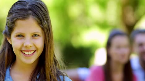 Little-girl-smiling-at-camera-with-parents-behind-on-park-bench