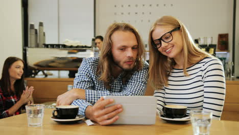 caucasian happy friends talking and watching a video on a tablet sitting at a table in a cafe