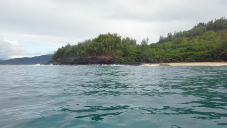 4K-Hawaii-Kauai-Boating-on-ocean-left-to-right-pan-from-mountains-in-distance-to-waves-crashing-on-rocky-shoreline-and-beach-with-clouds