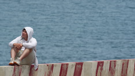 Asiatischer-Junger-Positiver-Mann,-Der-Auf-Einer-Strandbank-Sitzt-Und-Einen-Modernen-Kapuzenpullover-Vor-Dem-Meer-Trägt,-Sommerstimmung-Konzept