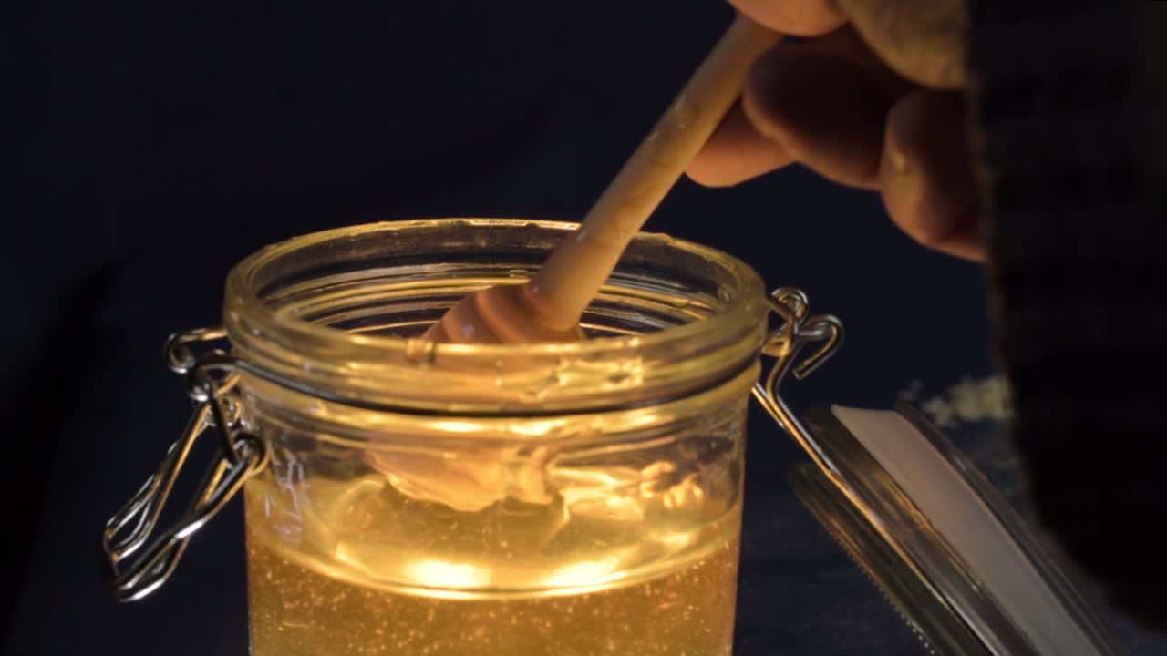 Mano che mescola miele dorato e liquido in un barattolo di vetro con cucchiaio dosatore, primo piano