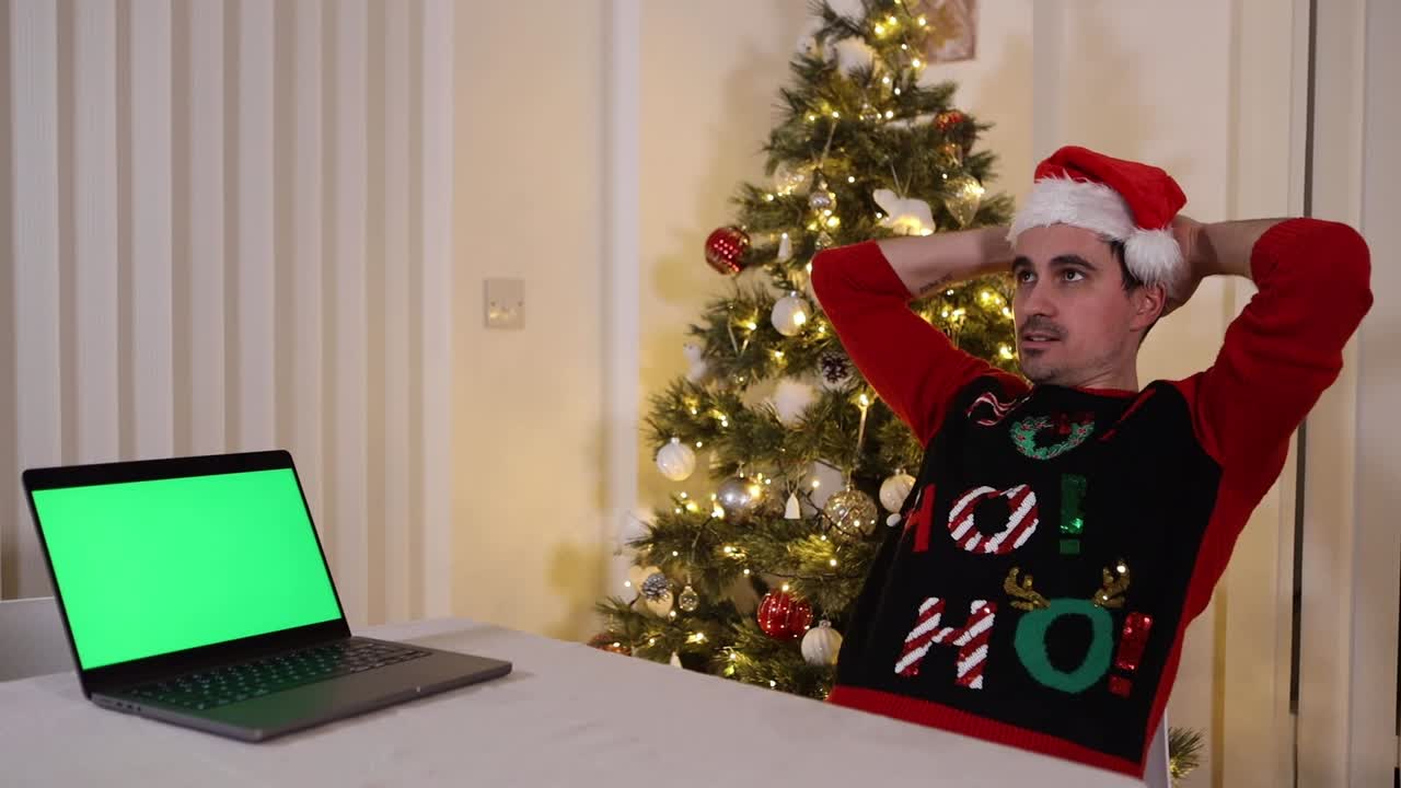 hombre navideño festivo viendo la pantalla verde del portátil cromakey en desacuerdo con lo que ve y recostándose en la silla con las manos en la cabeza pensando con expresión seria, frente a un árbol de navidad