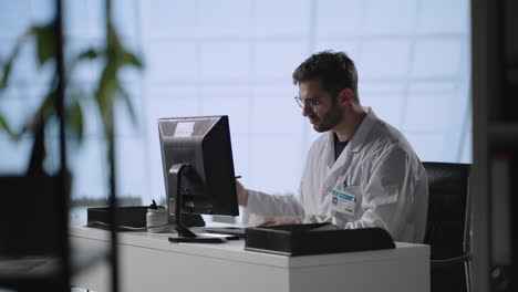 tilt down shot of male doctor in lab coat and glasses typing on laptop while working at desk in medical office