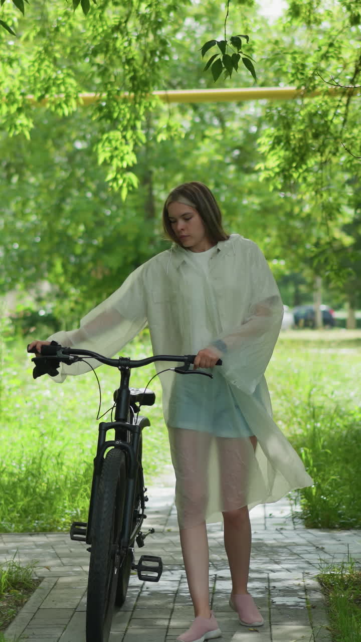 레인코트 를 입은 금발 여자 가 초록색 공원 을 가로지르는 포장 된 길 을 자전거 로 사려 깊게 산책 하고 있으며, 나무 와 풀 과 노란색 기둥 을 가진 도시 배경 으로 둘러싸여 있다
