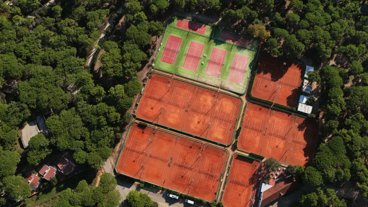 Vista aerea con drone di un grande complesso di tennis con diversi campi in terra battuta e campi verdi, circondato da una fitta foresta vicino alla costa