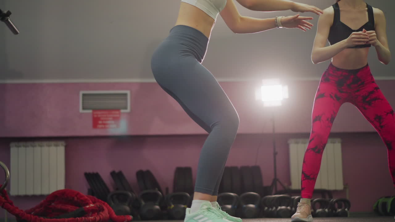 Istruttrice che guida una cliente negli squat in uno studio fitness, pareti rosa, manubri su rastrelliera, vista dal basso che enfatizza la spinta delle gambe e la forma, espressioni concentrate, retroilluminazione calda, ritmo controllato.