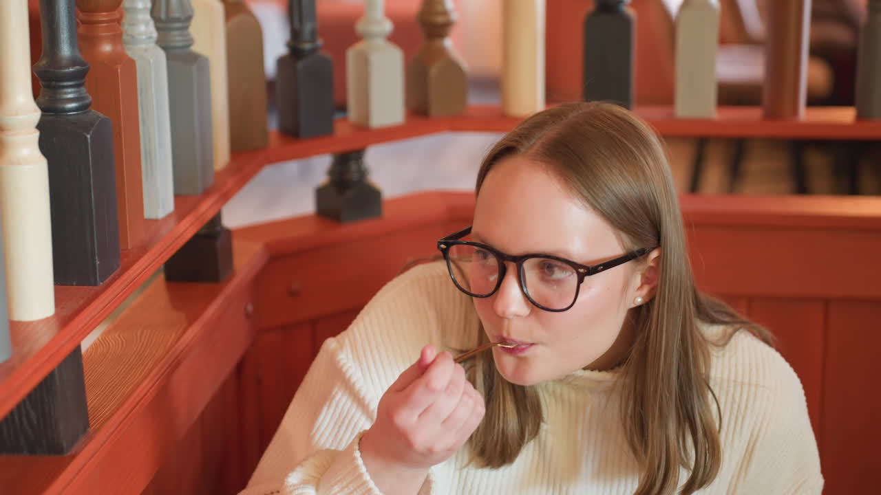 young woman in white sweater seated in cozy cafeteria, wearing glasses and looking down while enjoying chocolate cake, colorful wooden railing in background