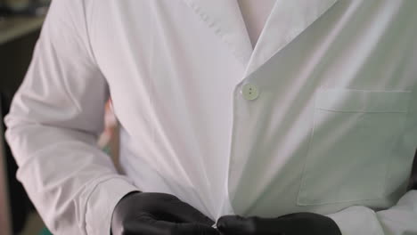 a close-up view of someone wearing black gloves as he carefully buttons his lab coat, with a view of a shelf behind